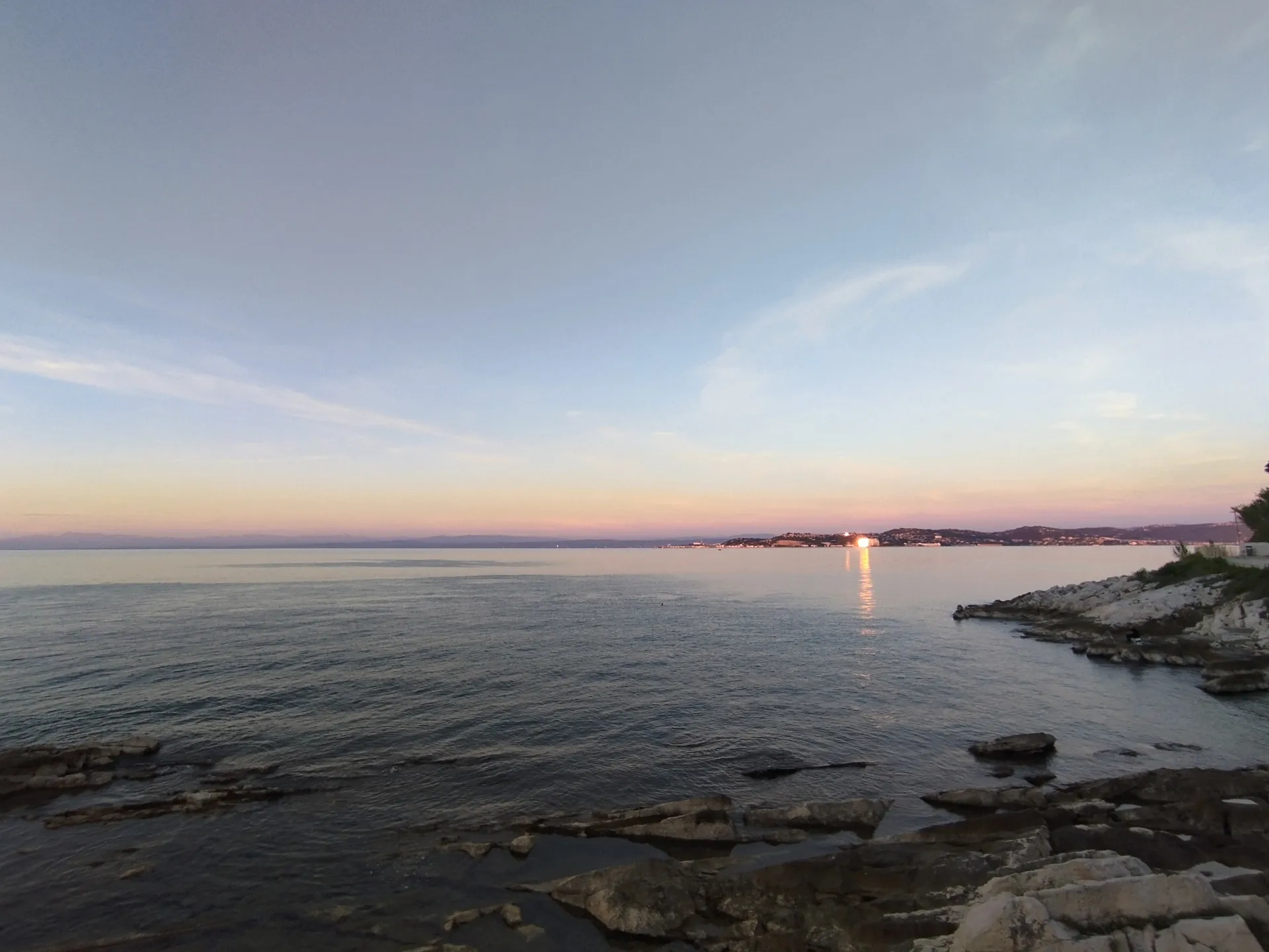 Calm water and flat limestone rocks at dusk, the lights of the Piran peninsula catching the last sun across the bay